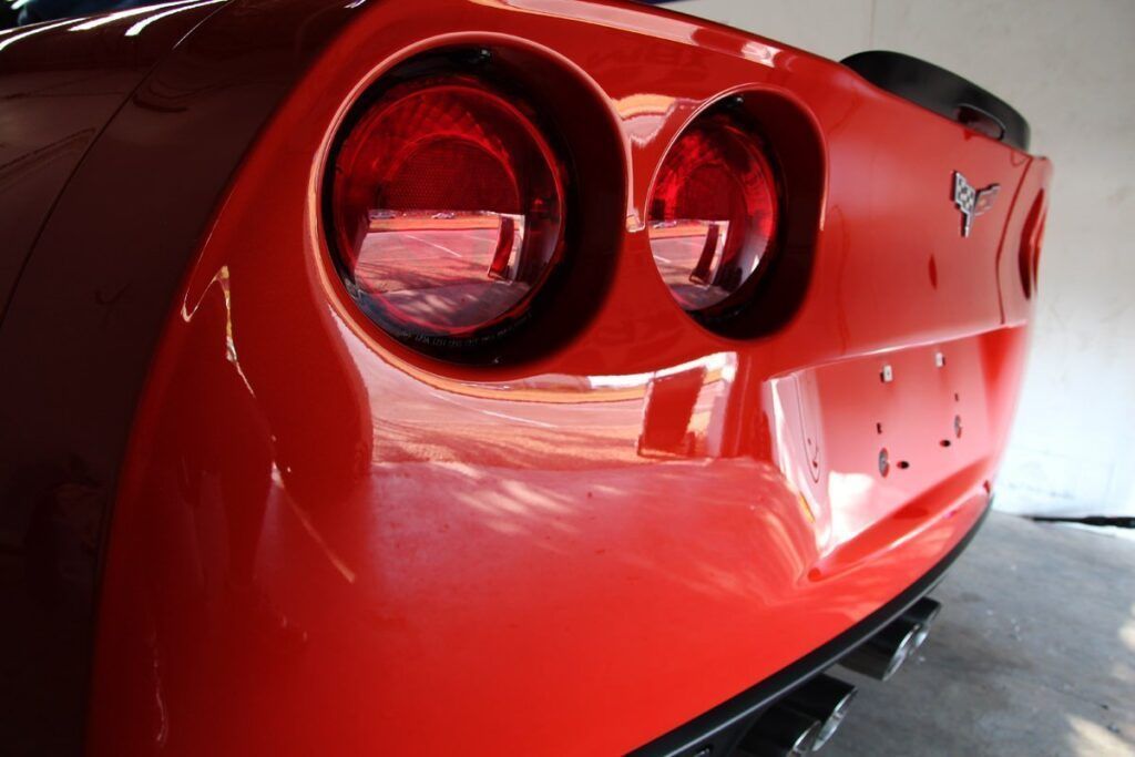 Red Corvette rear end, taillights illuminated. Shiny paint, outdoor setting.