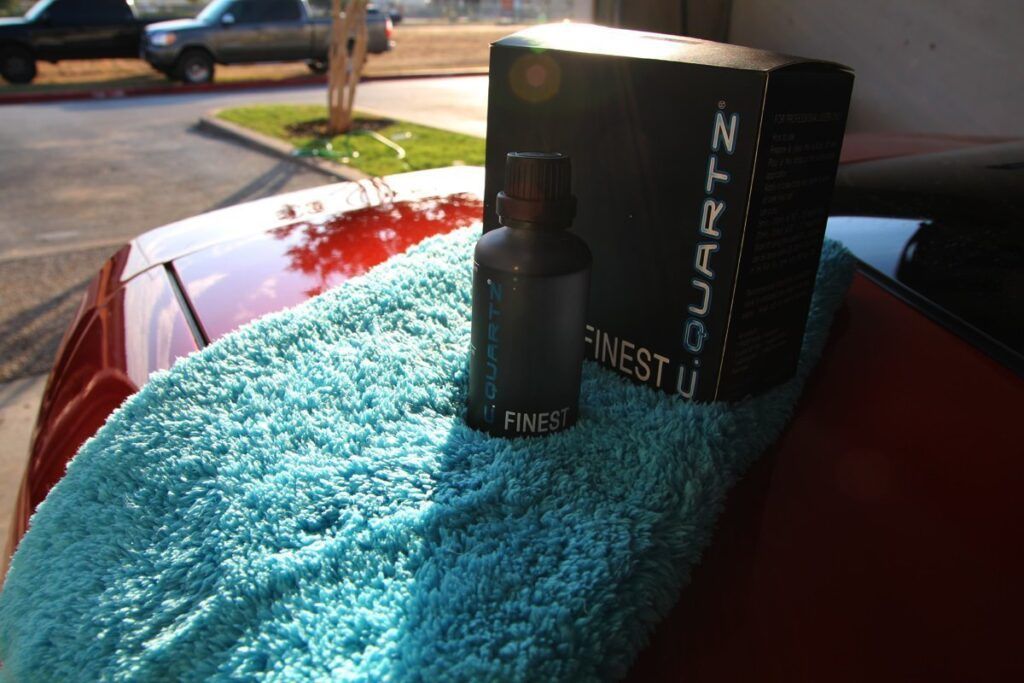 A bottle of car detailing product and its box on a blue towel atop a red car in daylight.