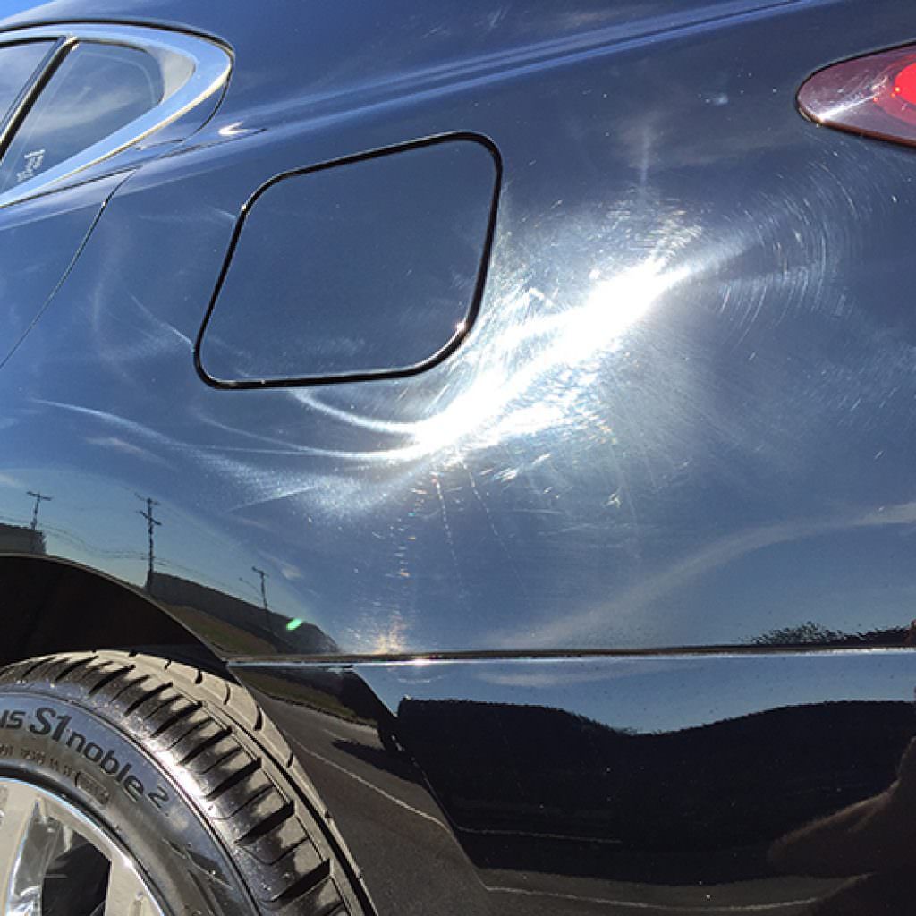 Black car body with visible scratches and swirl marks, reflecting sunlight and a blue sky.