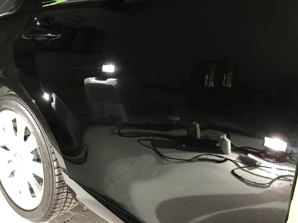 Black car's side with a shiny, reflective surface. Garage setting; shows reflections of lights and tools.