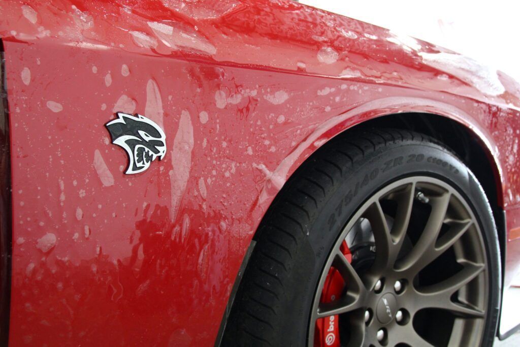 Red Dodge Challenger with Hellcat emblem on front fender, water droplets on the paint.