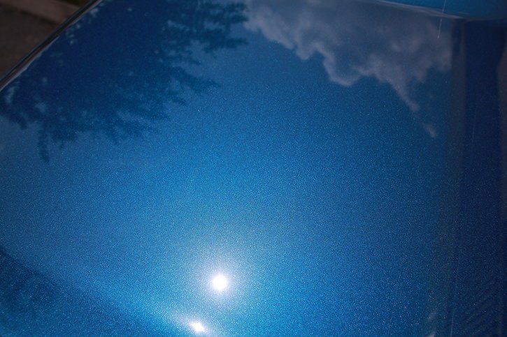 Blue car hood reflecting sky and trees, with sparkly metallic finish.