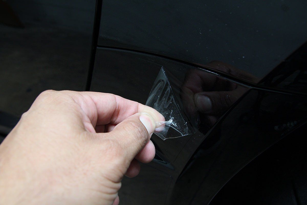 Hand peeling protective film from a black car's damaged surface, revealing scratches.