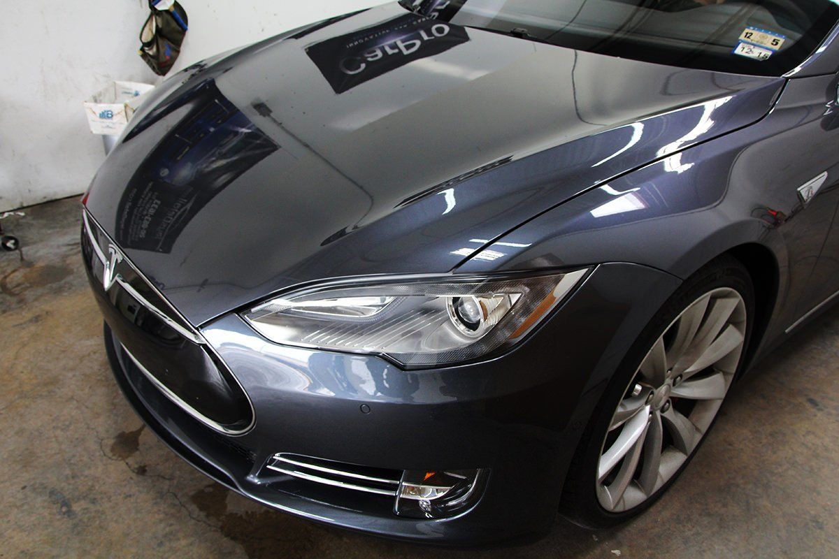 Dark gray Tesla car, front view, parked indoors, shiny paint, detailed headlights and wheels.