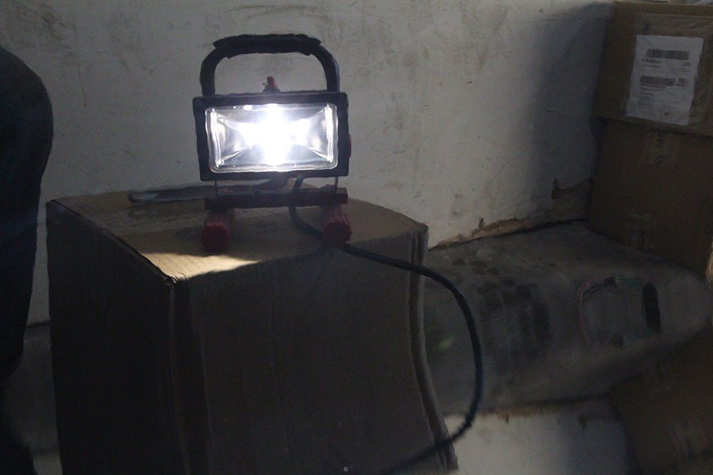 Bright work light on a box illuminates a dimly lit storage area. A black cord leads away.