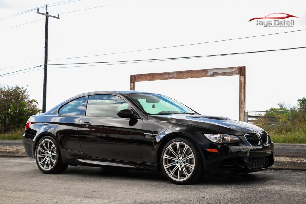 Black BMW M3 coupe parked on a street. Chrome wheels, sunny outdoors.