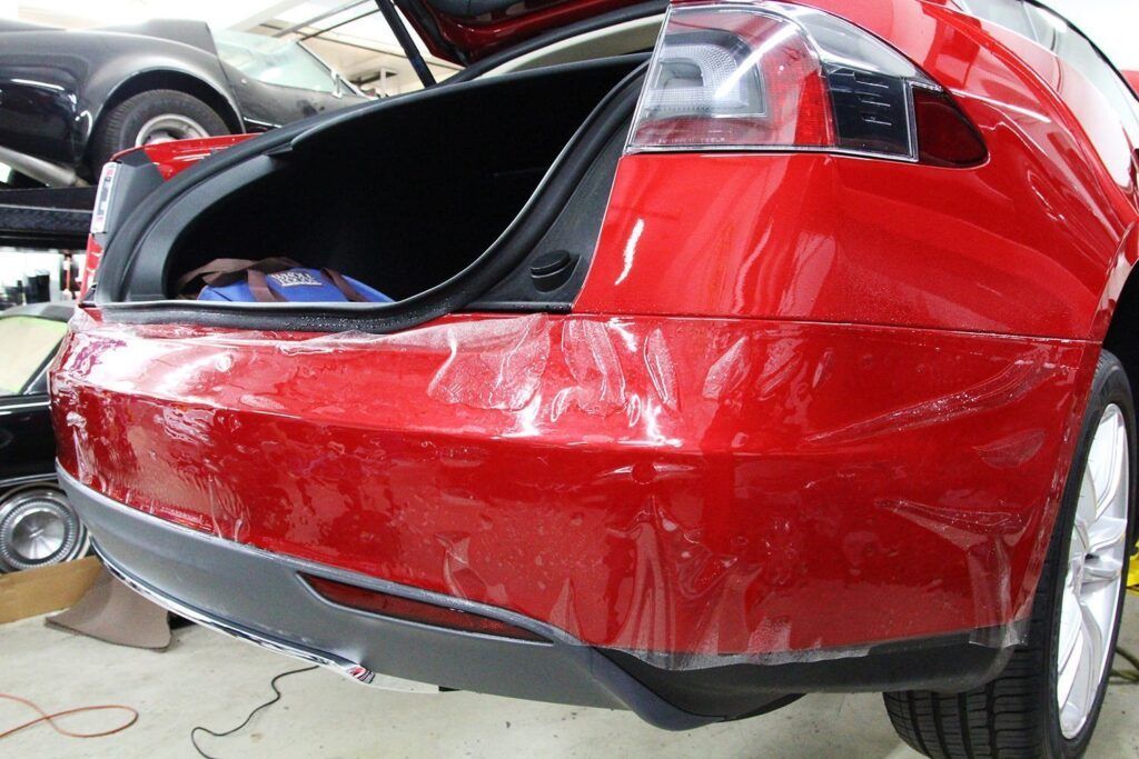Red Tesla with open trunk, plastic protective film on bumper, in a garage.