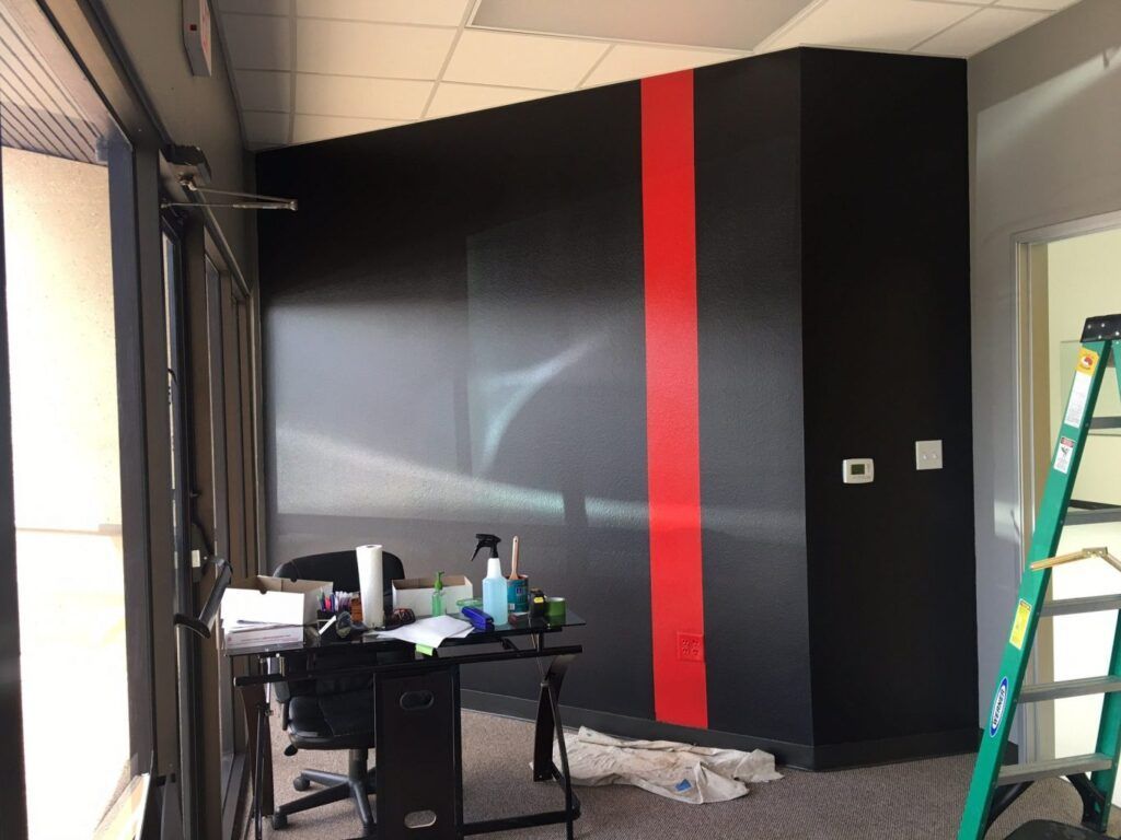 Black wall with red stripe; office setting with desk, window, and ladder.