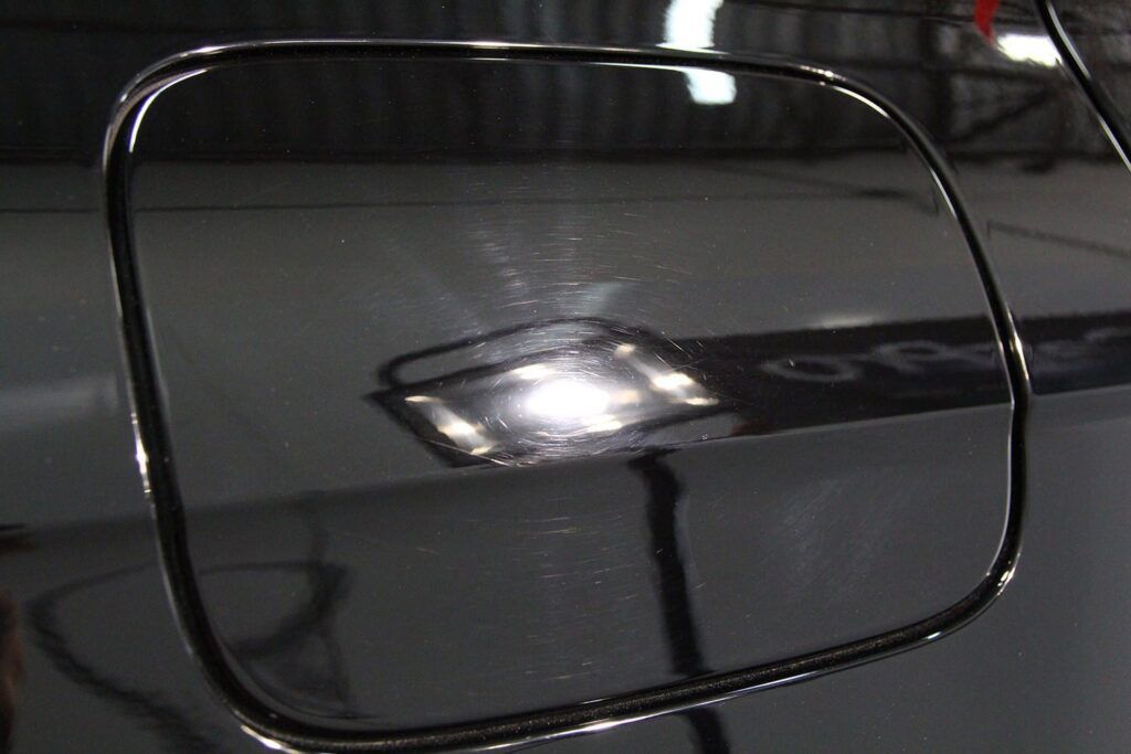 Black car fuel door with scratches, reflecting overhead lights.