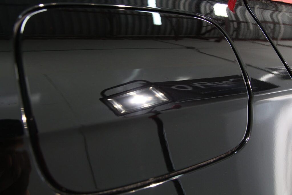 Close-up of a shiny, black car fuel door reflecting a bright light.