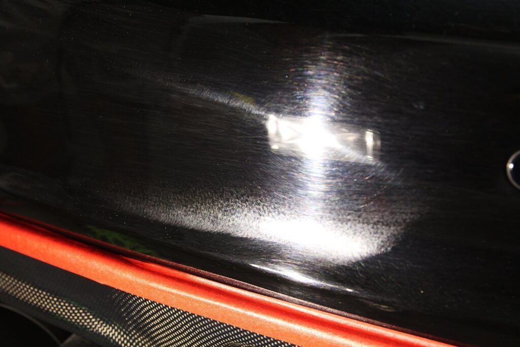 Black car panel with swirls reflecting light, above red and carbon fiber trim.