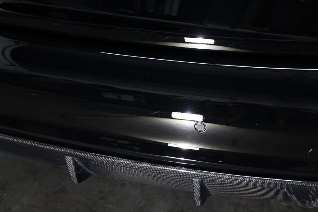 Close-up of a black car bumper with carbon fiber trim, reflecting light.