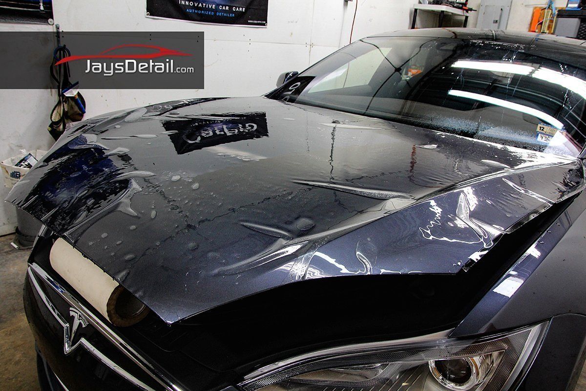 A person is applying a protective film on the hood of a Tesla. The car is dark gray.