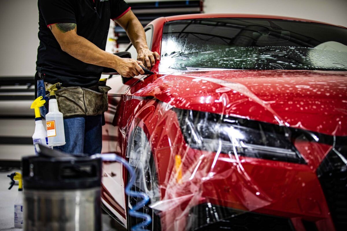 Man applying protective film to a red car in a shop.
