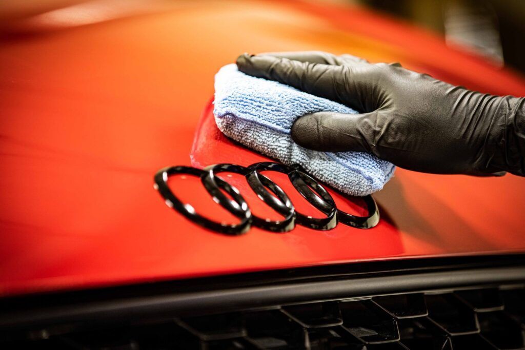 Gloved hand using a blue cloth to polish a red Audi emblem on a red car hood.