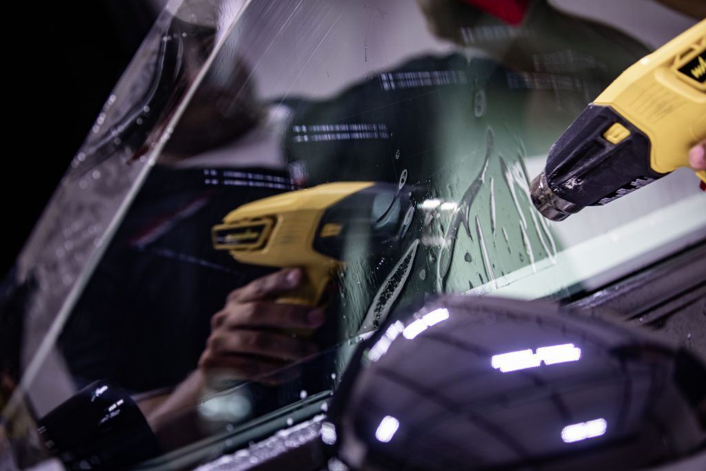 A person uses a heat gun to apply tinted film to a car window, likely in a garage.