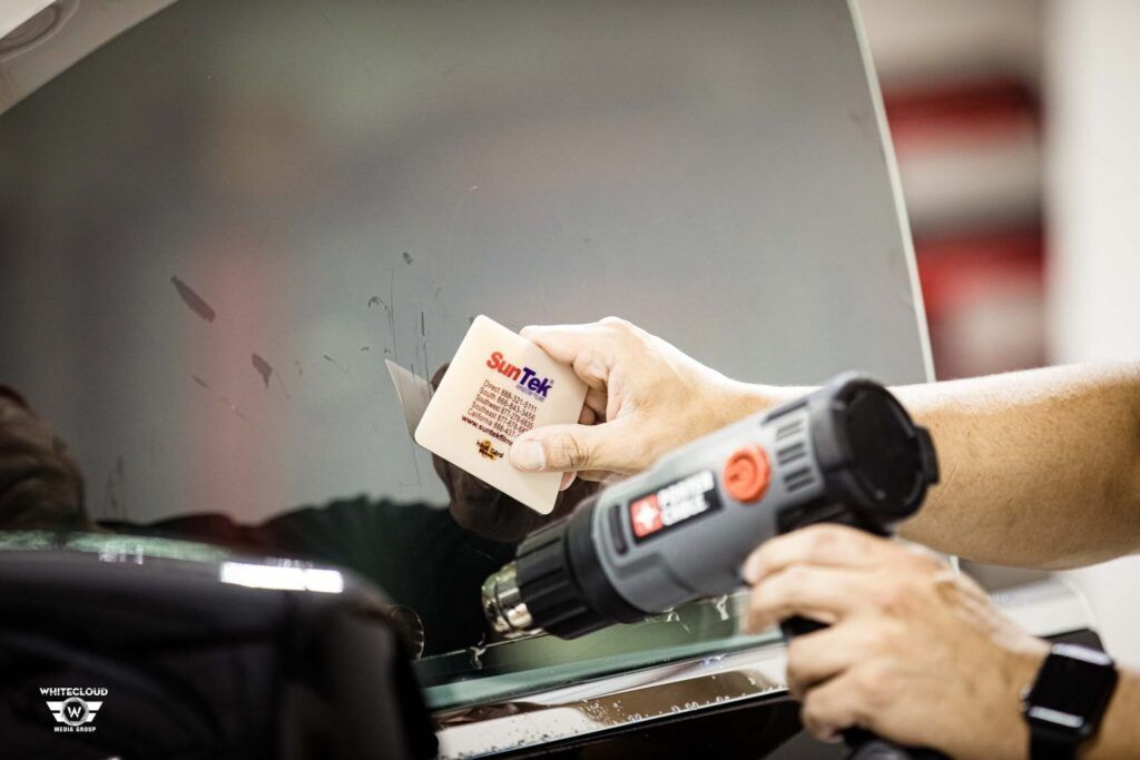 Person applying tint to a car window using a heat gun and a squeegee in a shop.