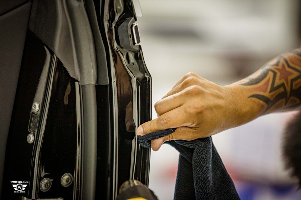 Person's hand with tattoos wiping a black car door with a cloth in a garage.