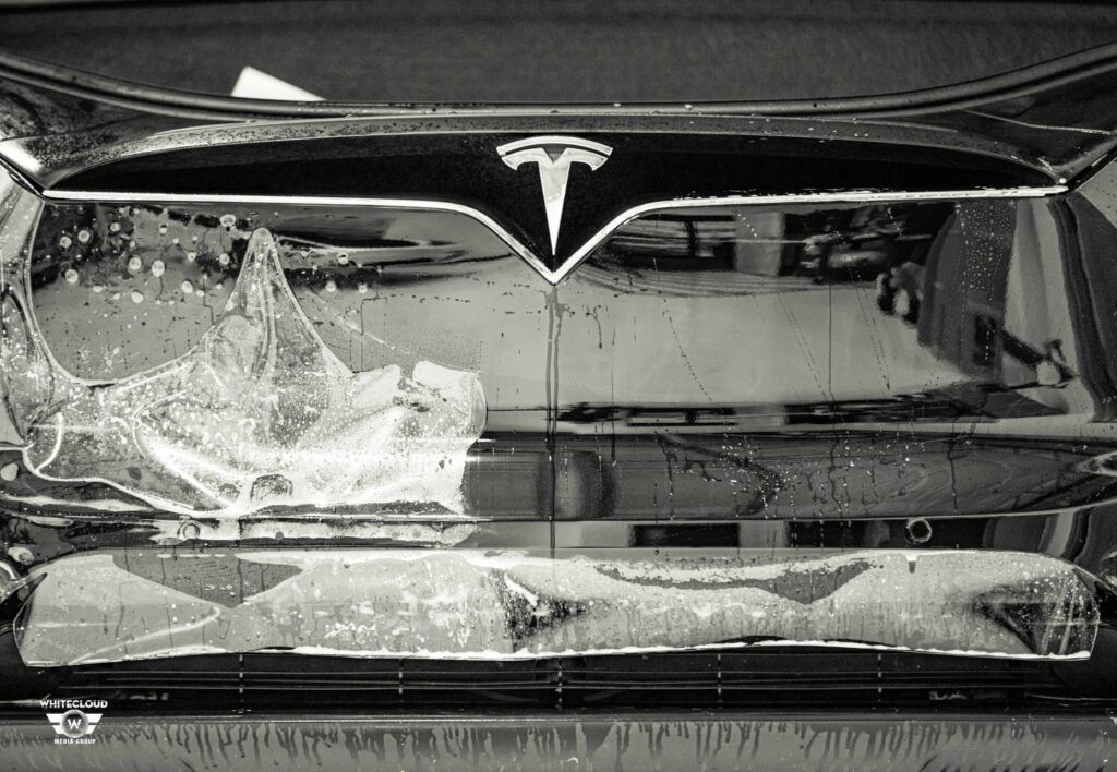Black and white close-up of a damaged Tesla car's front. Windshield shattered, Tesla logo prominent.