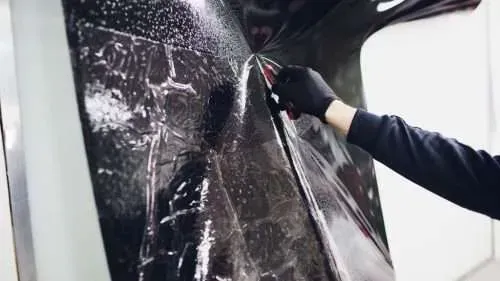 A person with a gloved hand is trimming dark film on a surface with a knife.