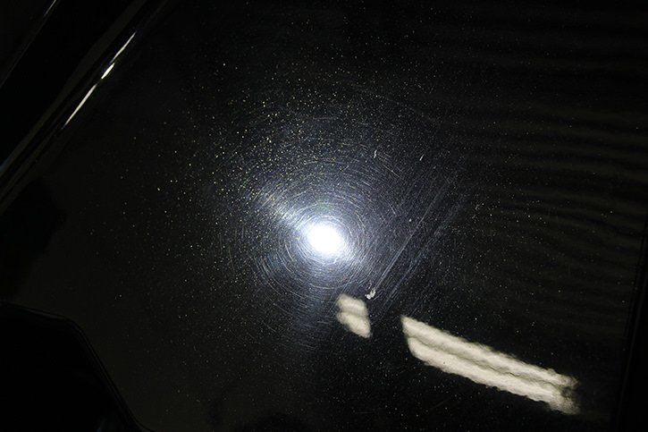Close-up of shiny, black car paint surface with swirl marks and a bright reflection.