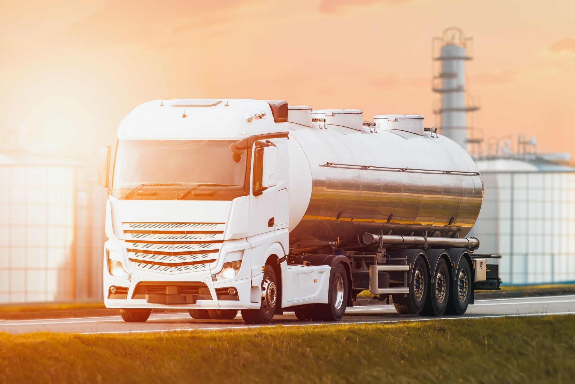 White tanker truck driving past industrial tanks at sunset on a road. White tanker truck driving past industrial tanks at sunset on a road.
