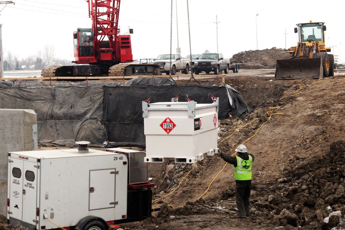 Crane lifting a white fuel tank at a construction site with worker in safety vest.