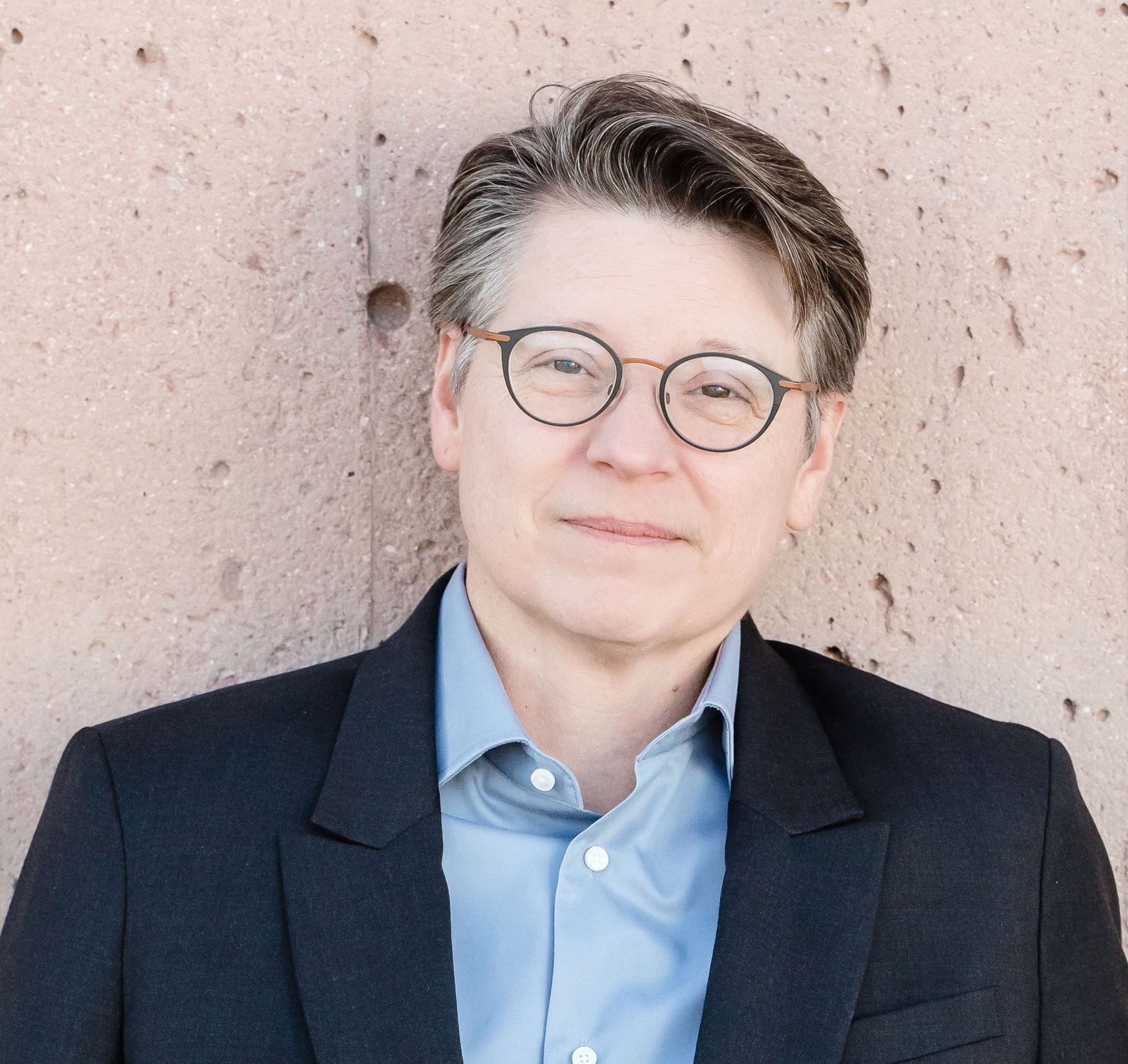 Person in glasses and blue shirt, wearing a dark blazer, standing against a textured wall.