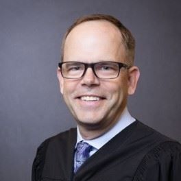 Portrait of a smiling man in glasses wearing a black suit and purple tie against a gray background