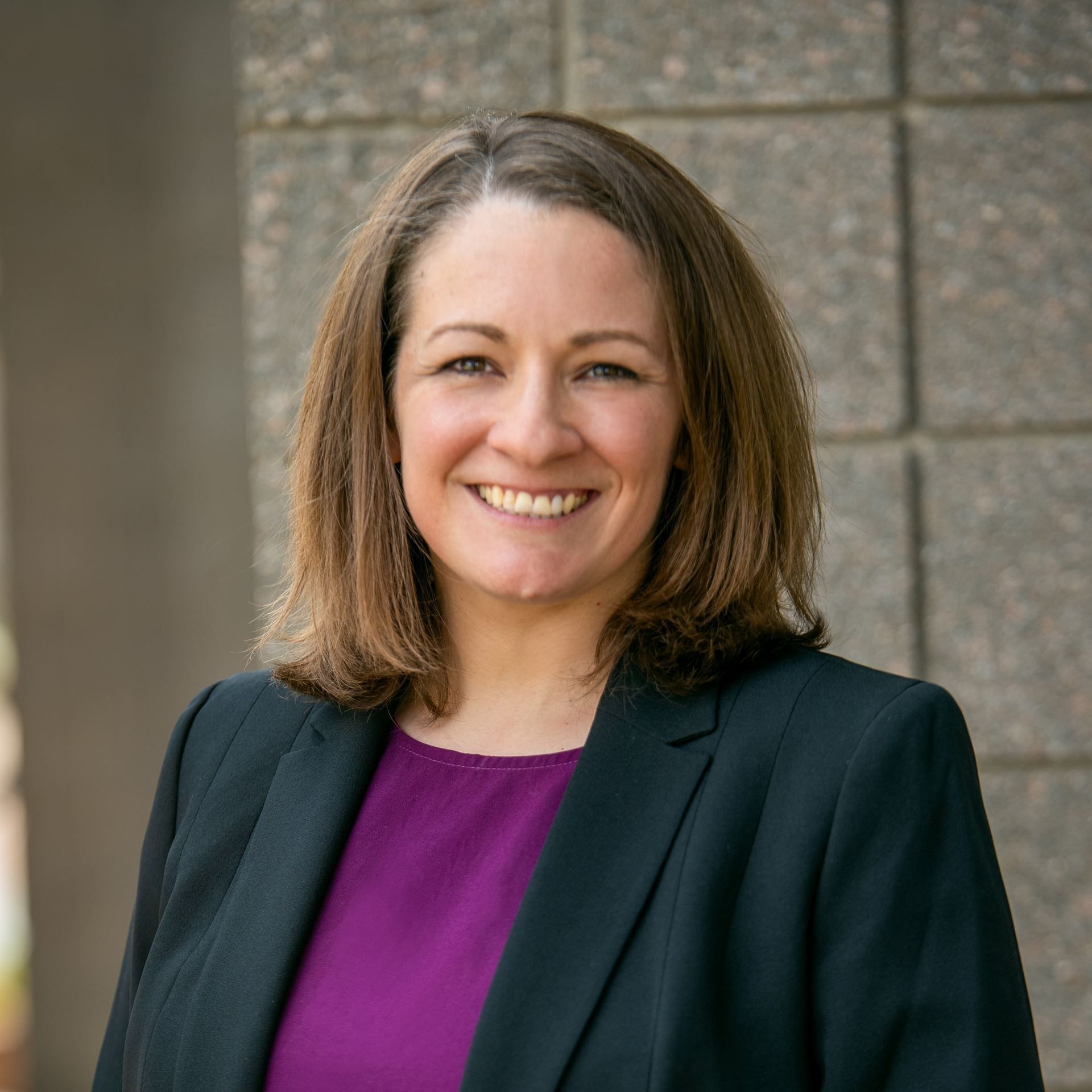 Smiling person in a black blazer and purple top standing before a stone wall.