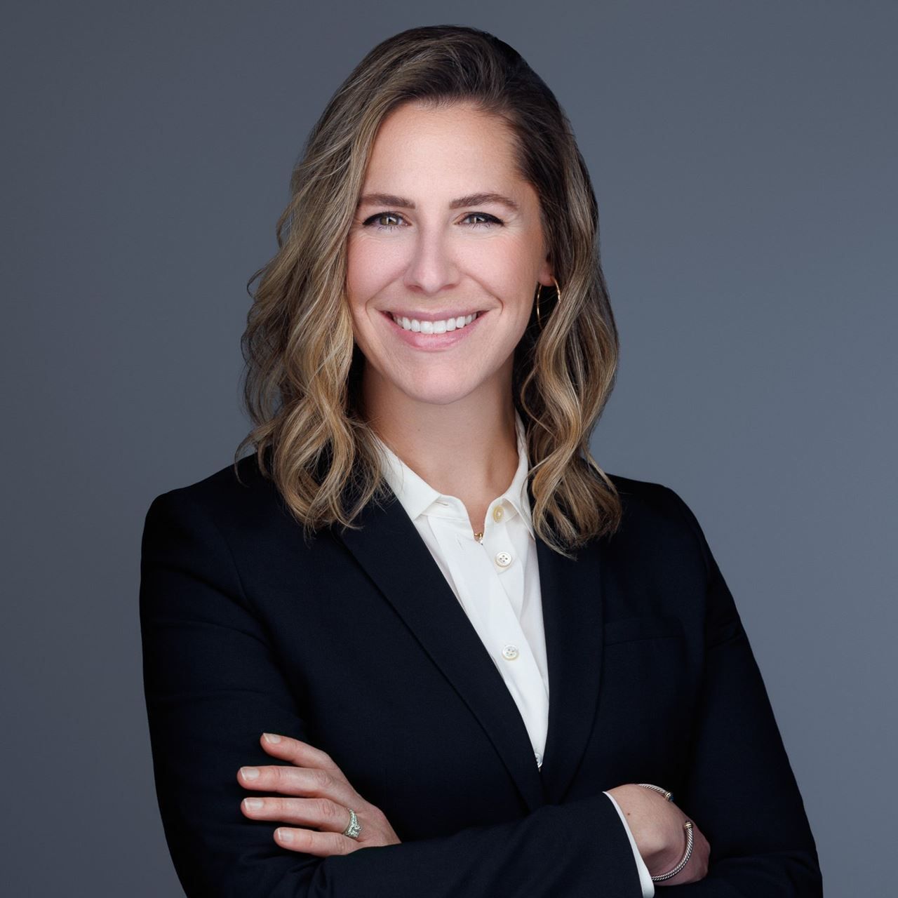 Professional headshot of a smiling person in a black blazer and white shirt on a gray background