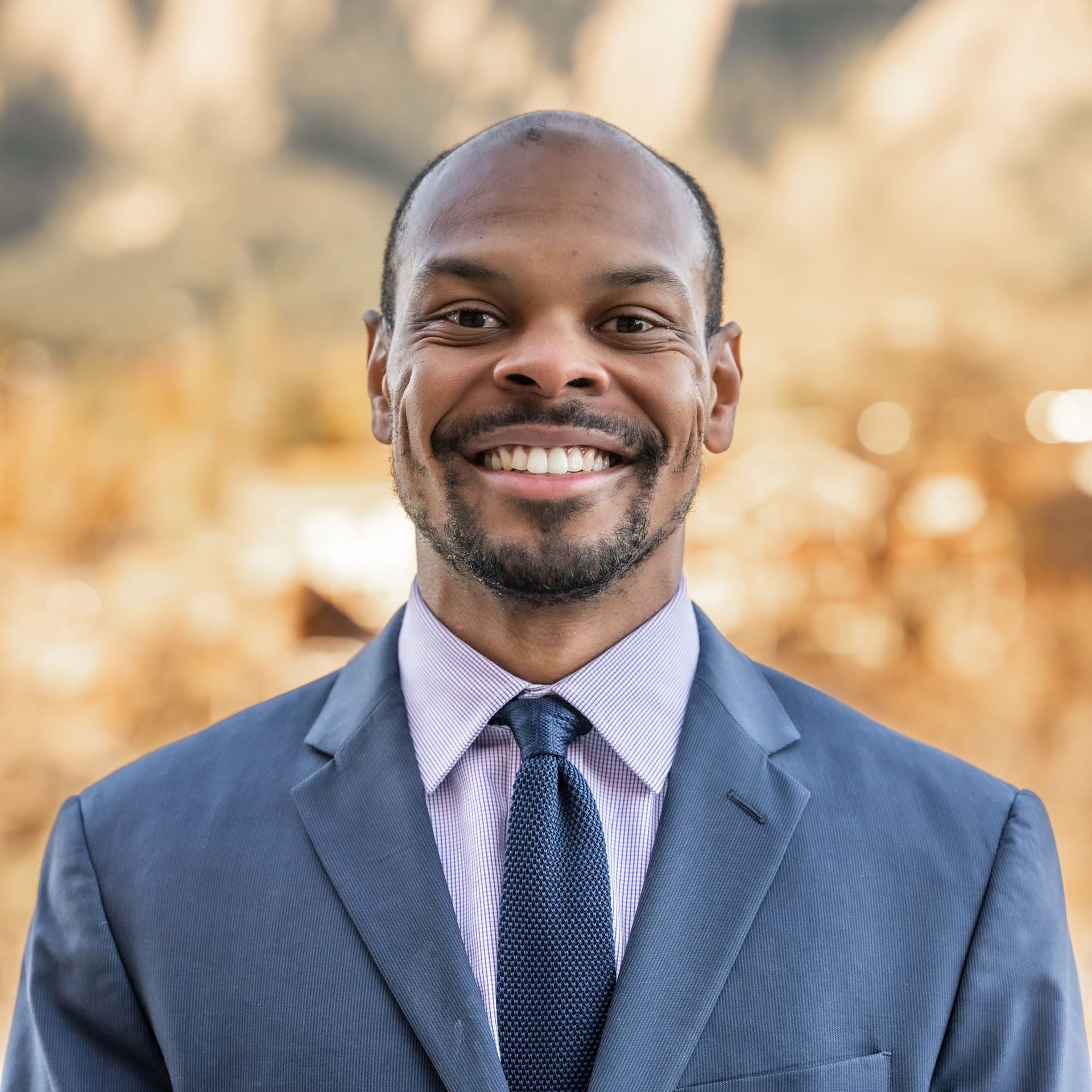 Smiling man in a blue suit and tie outdoors with blurred trees in the background