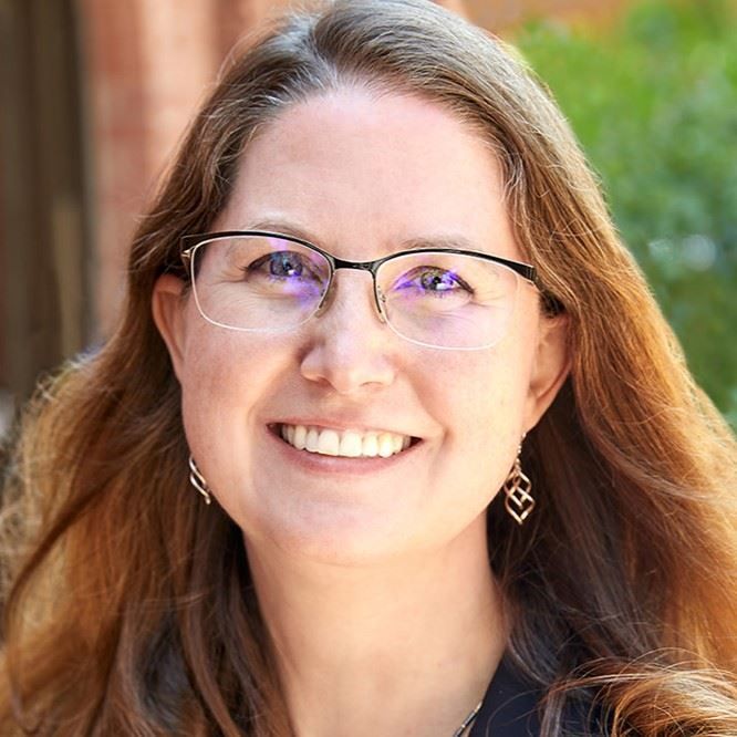 Smiling person with glasses and long hair outdoors against a blurred brick background