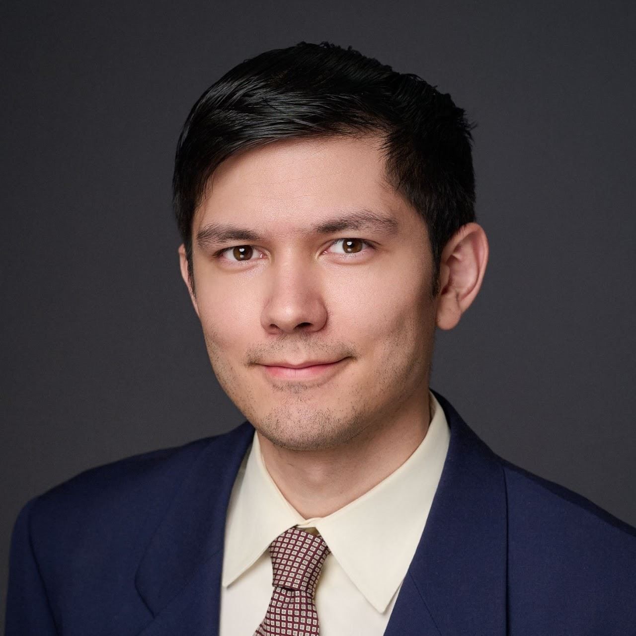 Studio headshot of a person in a navy suit and patterned tie against a dark gray background