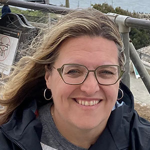 Smiling person wearing glasses and hoop earrings outdoors on a windy day with a metal railing behind them