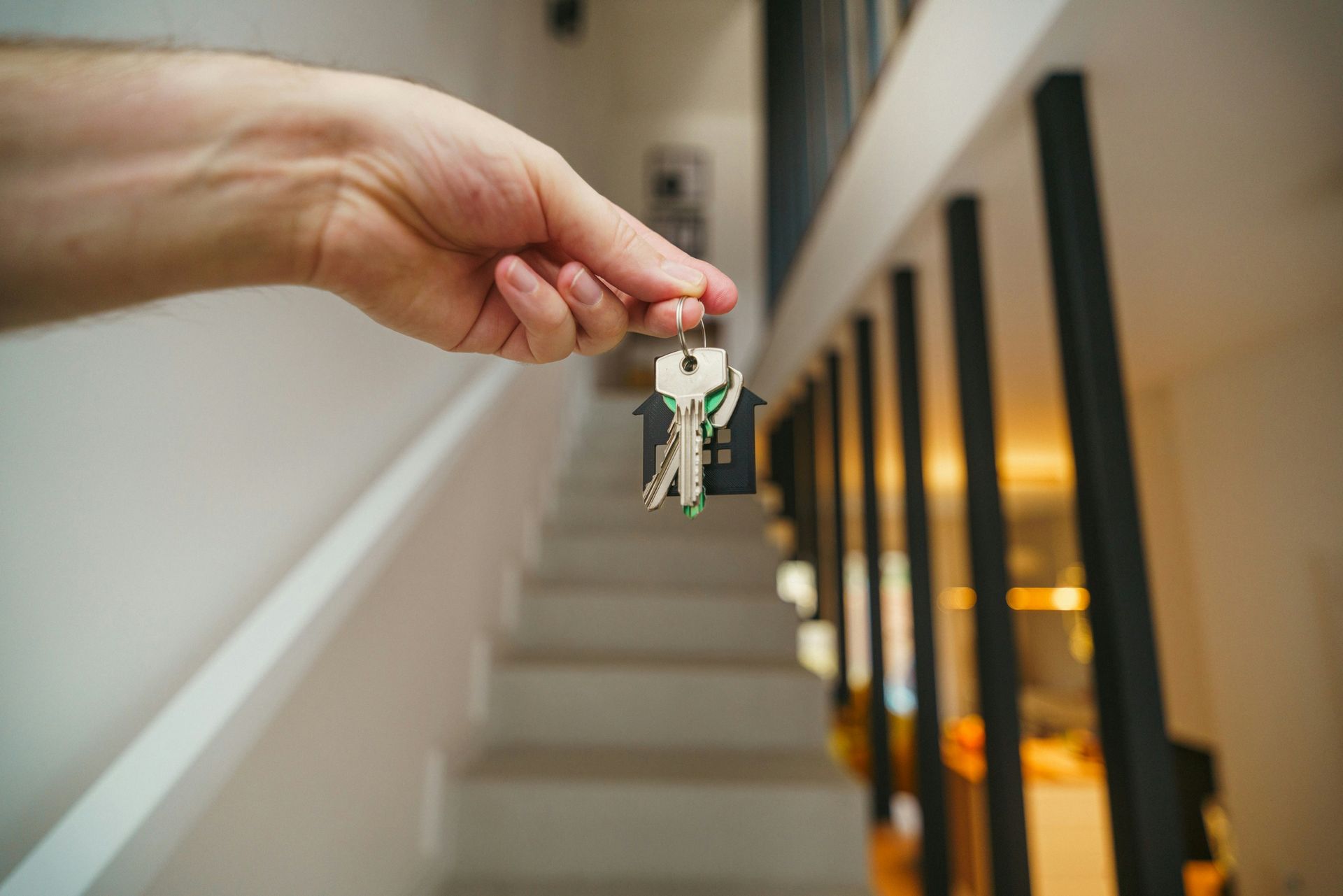 A hand holding a house-shaped keychain with keys, positioned in front of a modern staircase inside a home.