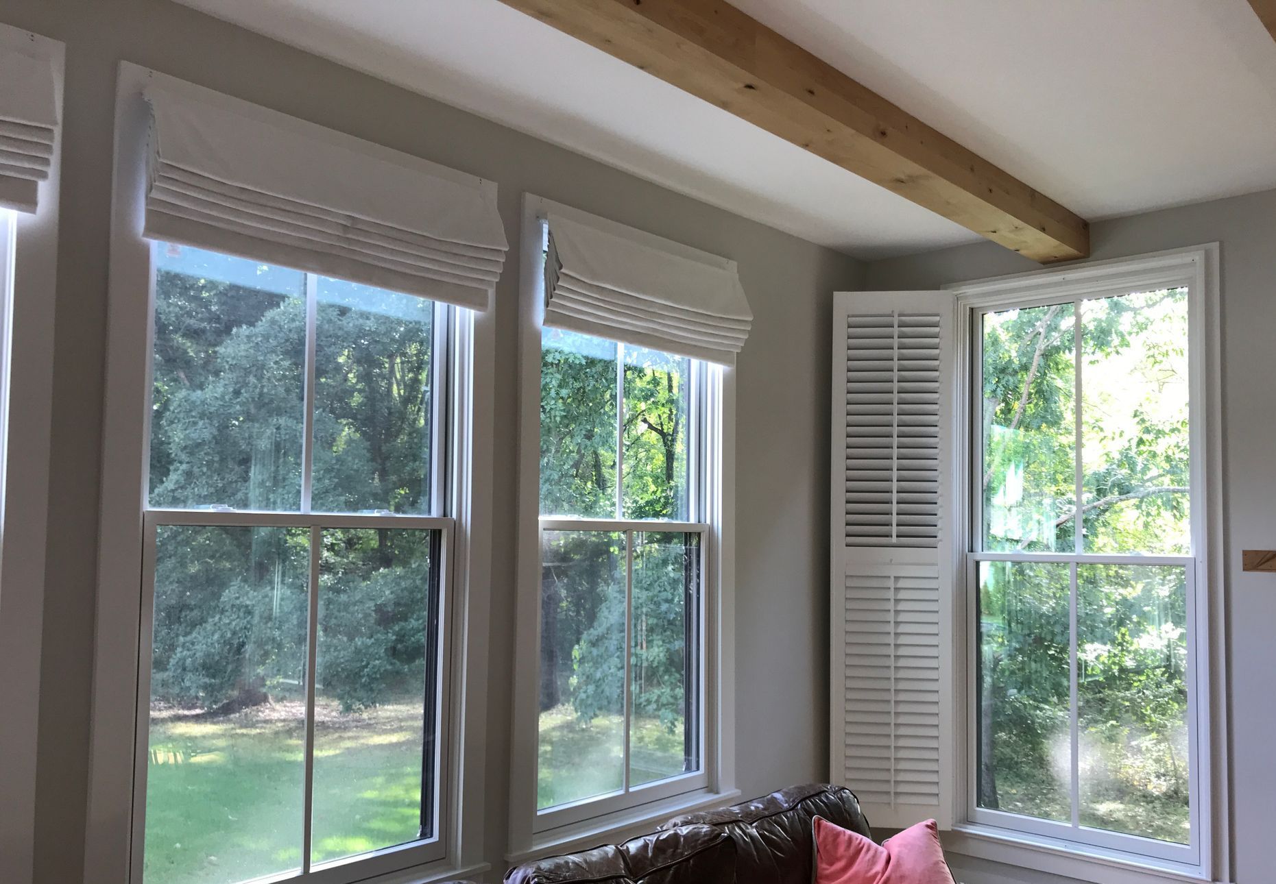 Three craftsman-style windows are visible in a room with light gray walls. Two windows on the left have white roman shades pulled up, revealing a view of green trees outside.