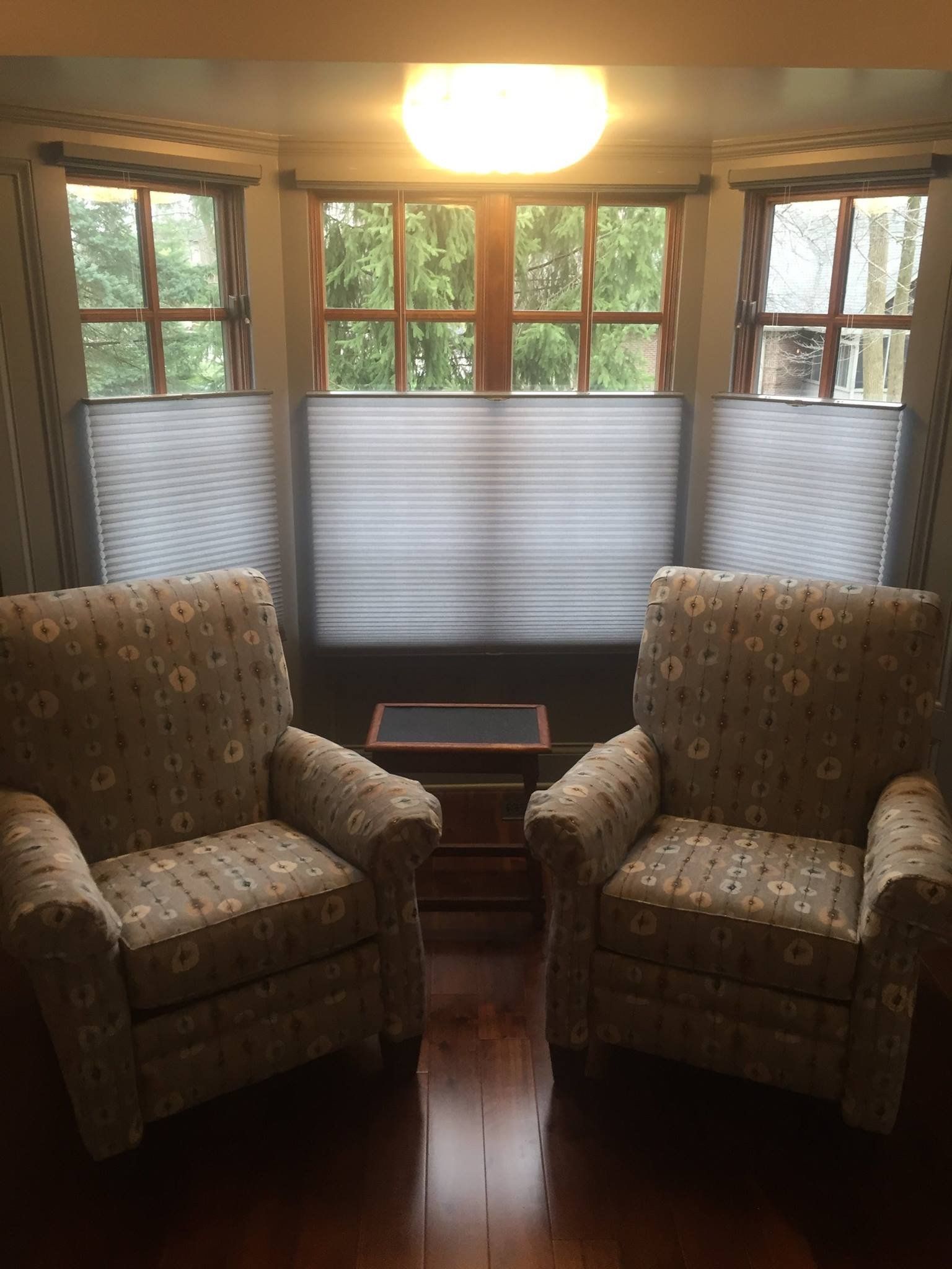 Three windows, likely part of a bay window, are fitted with light-colored cellular shades in a top-down, bottom-up style. The cellular shades are lowered to cover the bottom half of each window. Outside, green trees are visible through the uncovered upper portions of the windows. In front of the windows, two upholstered armchairs with a floral pattern are positioned, facing away from the viewer, with a small end table between them. A warm light fixture is visible on the ceiling above the window.