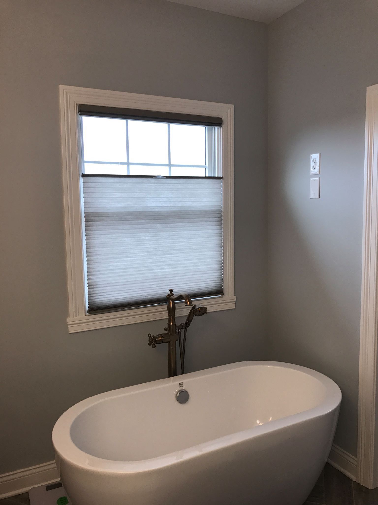 A freestanding white bathtub sits beneath a window fitted with light gray cellular shades. The cellular shades are installed in a top-down, bottom-up style, with the top portion of the window revealing a bright sky. A bronze-toned faucet with a handheld shower attachment extends from the floor into the tub. The bathroom walls are painted a light gray, and white trim outlines the window and doorway.