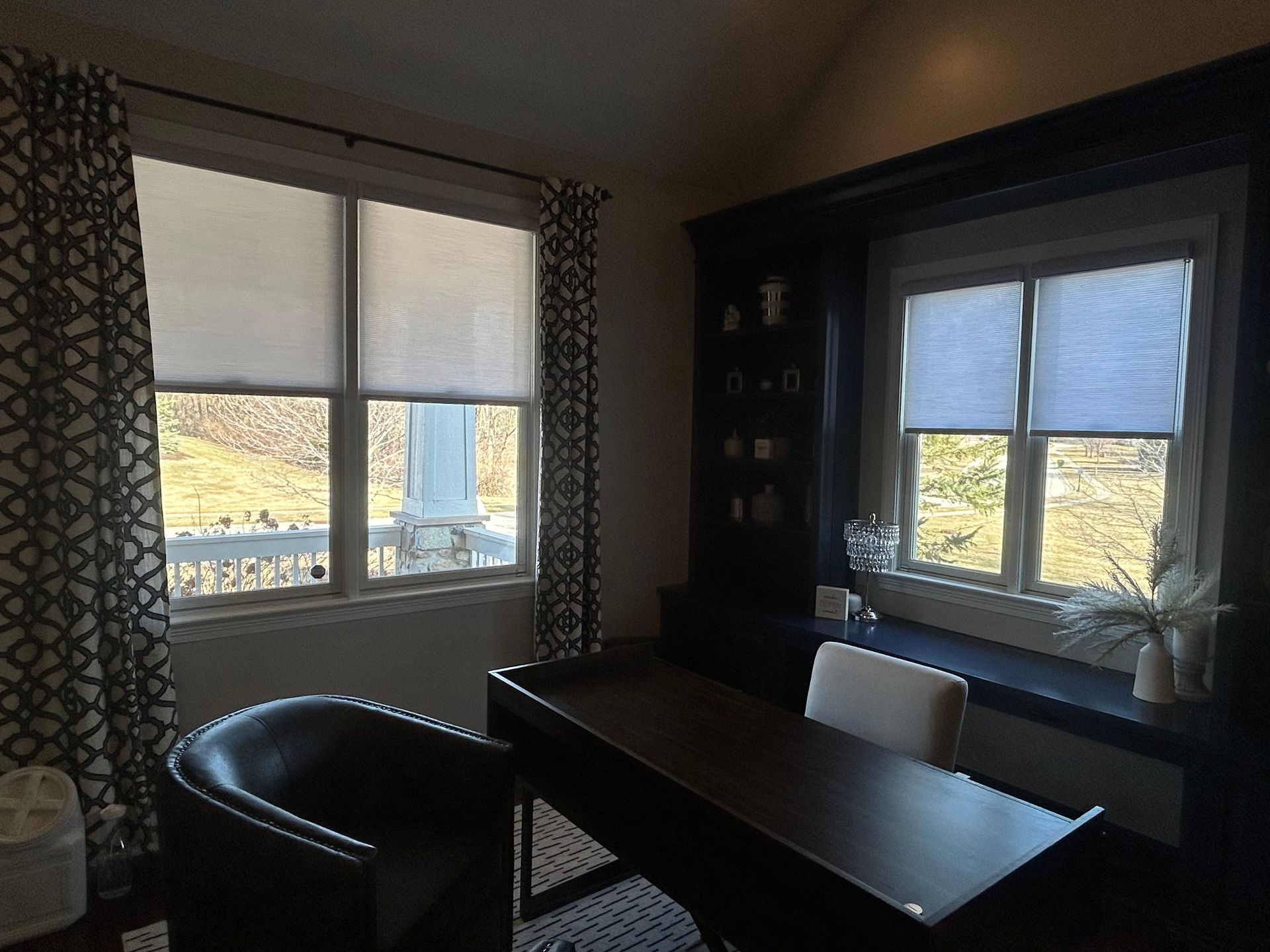 An interior shot captures a home office space. Two large windows on the left, partially covered by patterned curtains and white roller shades, allow natural light to filter into the room and offer a view of the outdoors. A dark wooden desk dominates the foreground, paired with a black leather chair tucked underneath and a white chair positioned on the opposite side. Built-in dark blue cabinetry lines the far wall, featuring shelves adorned with decorative items and a section that serves as a window seat or another work surface with a lamp and vases. The overall ambiance is that of a functional yet stylish workspace within a home.