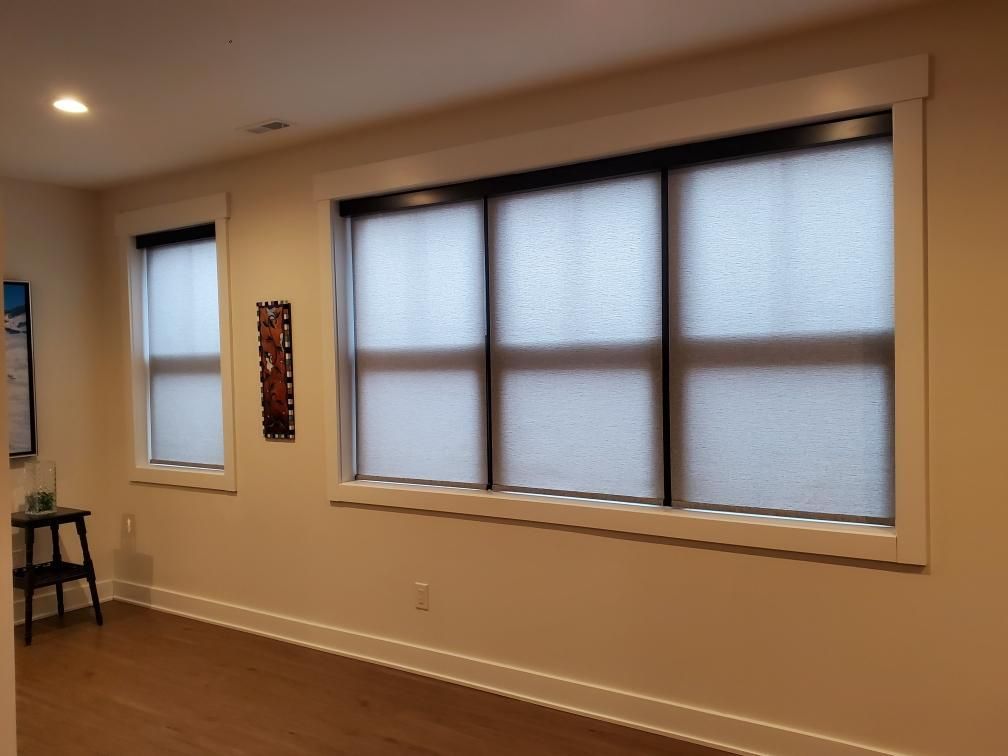 A warmly lit room with beige walls and a dark wood floor features two windows fitted with dark-framed roller shades. The larger window on the right has three separate roller shades covering it, while the smaller window to the left has a single roller shade.