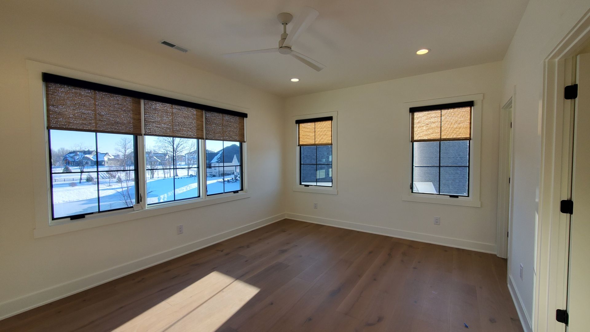 A well-lit room with light beige walls and warm-toned hardwood flooring. Three windows are visible, each fitted with dark-framed roller shades made of a woven natural material. The largest window shows a snow-covered landscape outside, revealing houses and bare trees under a bright sky. Two smaller windows on the adjacent wall also have the same type of roller shades and offer a view of the outdoors. A white ceiling fan with four blades is mounted on the ceiling. Two recessed lights provide additional illumination. A partially visible white door frame is on the right side of the image.
