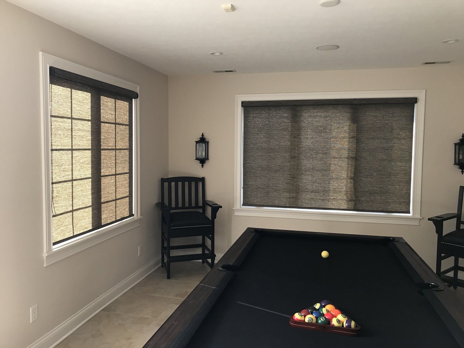 A game room with light beige walls and a light-colored tiled floor features two windows covered with dark-framed roller shades made of a woven material. The window on the left is taller and narrower, while the window on the right is wider and shorter. A black pool table with a black felt surface dominates the foreground, with a set of billiard balls racked at one end and a single yellow ball near the center. Two black chairs with padded seats are positioned against the wall, one under a decorative wall sconce. The room is illuminated by recessed lighting in the ceiling.