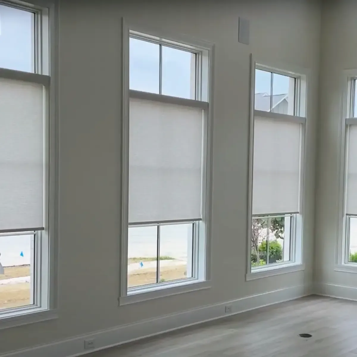 Four tall, white-framed windows are visible in a room with light-colored walls and light wood flooring. Each window is equipped with light-filtering motorized shades that are partially lowered, covering the top portion of the window.