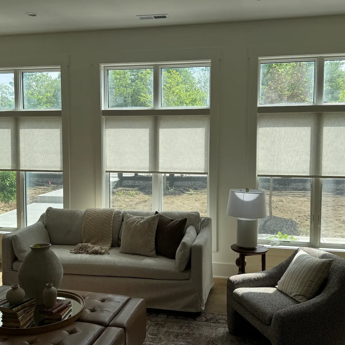 A well-lit living room features three large windows with light-colored roller shades covering their upper half, allowing sunlight to stream in. 