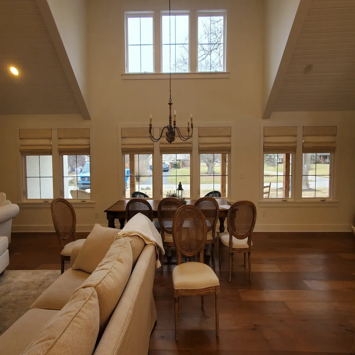 A warmly lit living and dining area features a wooden dining table surrounded by six chairs with oval backs and light-colored cushions. Natural light streams in through multiple windows fitted with light-colored roman shades. A traditional chandelier hangs above the dining table. In the foreground, a portion of a light-colored sofa with throw pillows and a blanket is visible on a rug. The room has high ceilings and hardwood floors.
