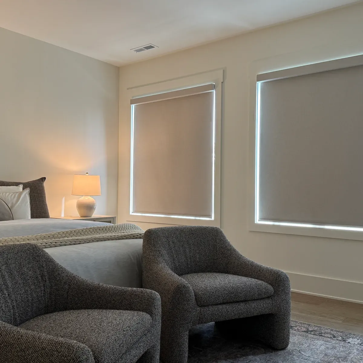 A cozy bedroom features two armchairs in the foreground, with room darkening shades on two large windows in the background. A bed with a lamp on a nightstand is partially visible on the left.