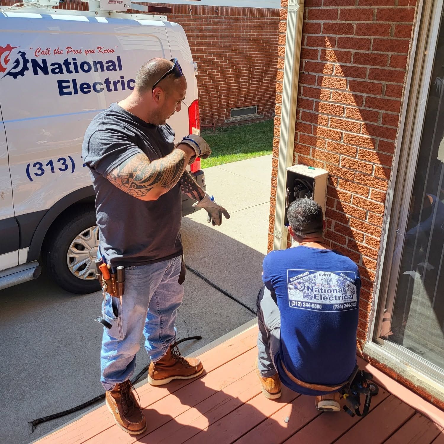 Two workers on a porch beside a National Electric van, inspecting or repairing wiring near a brick house.