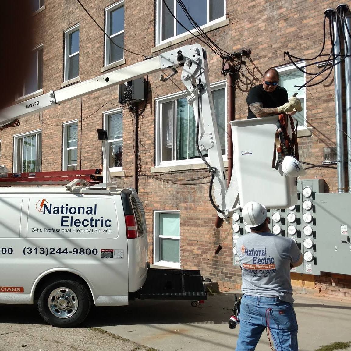 Electric utility workers in bucket trucks repairing overhead lines beside a brick building.