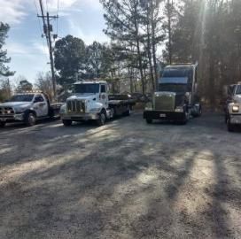 Four tow trucks parked on gravel, trees and power lines in background, sunlight.